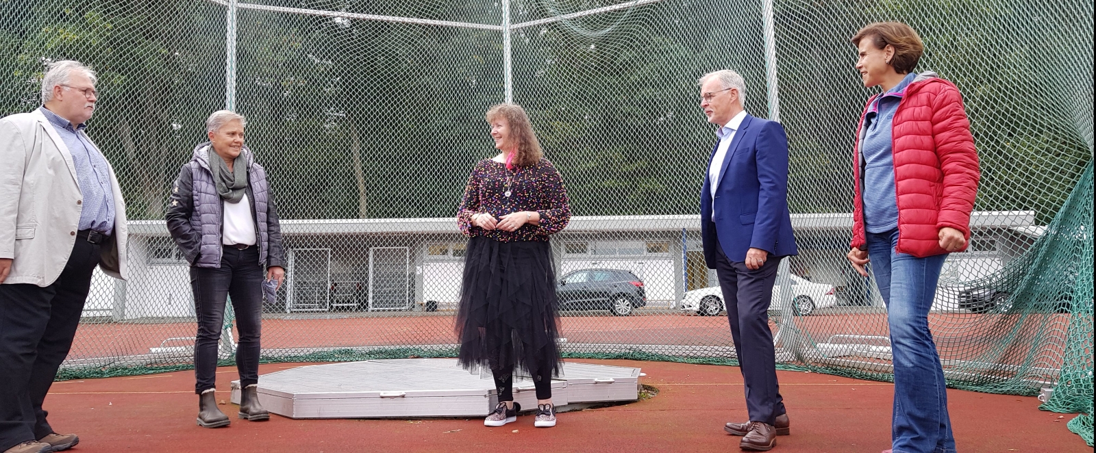 Staatssekretärin Andrea Milz steht mit mehreren Vereinsmitgliedern auf einem Hammerwurf-Sportplatz. Im Hintergrund sieht man das Auffang-Stahlnetz im Halbkreis aufgestellt.