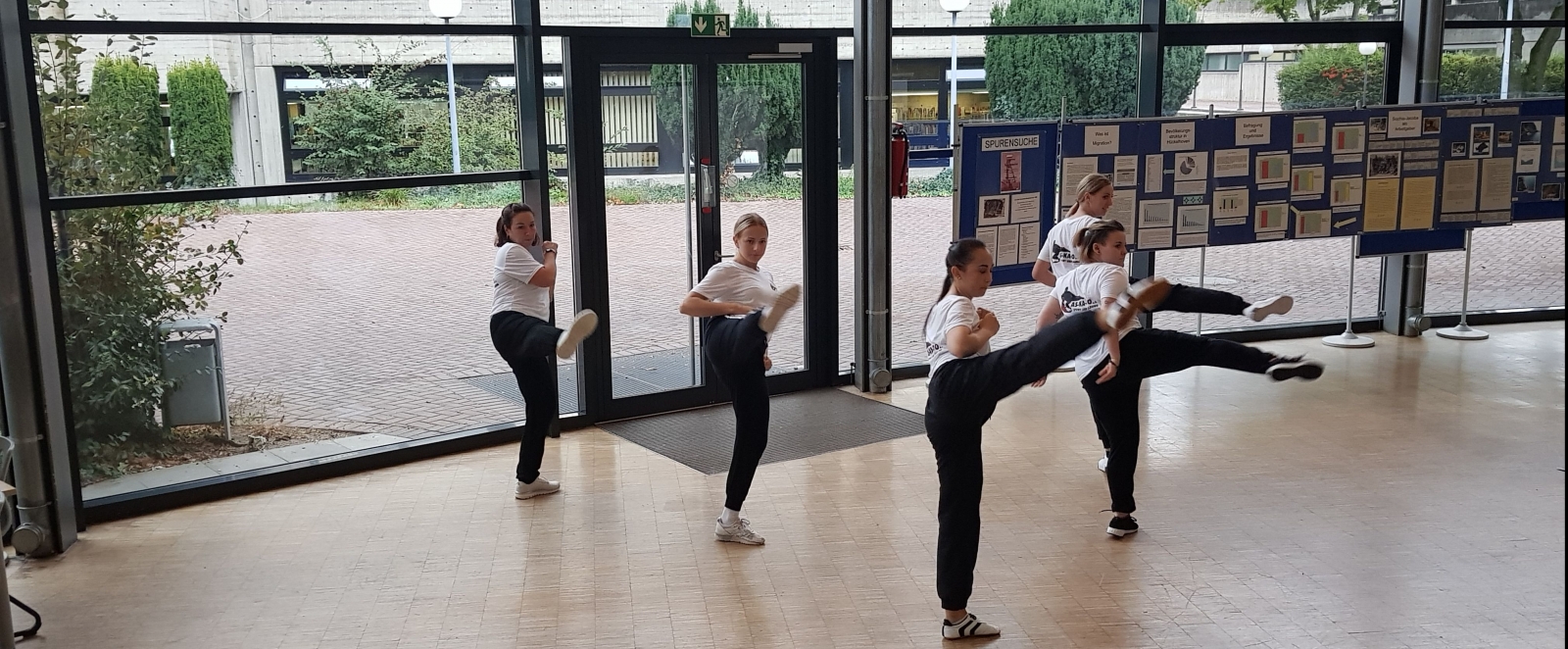 Fünf junge Damen in schwarzen Sporthosen und weißen T-Shirts demostrieren Selbstverteidigung und machen alle zusammen mit ihren rechten Bein einen Seitwärts-Tritt.