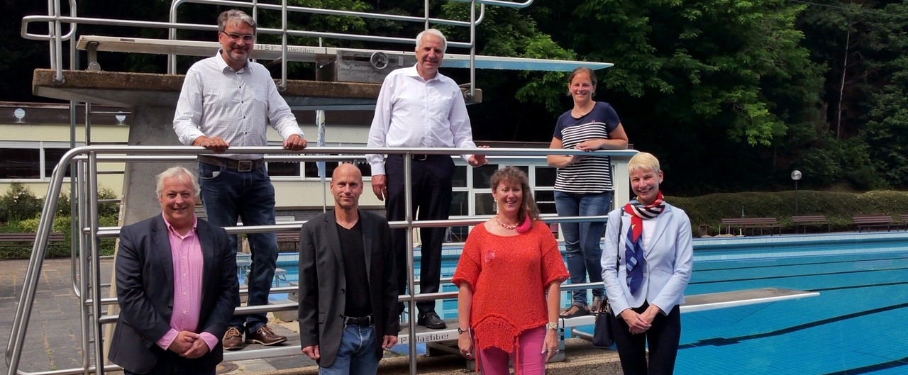 Staatssekretärin MIlz im rot-orangem Outfit an einem Freibad, hinter ihr sind zwei Springbretter zu sehen. Mit ihr sind noch sechs weitere Personen vom Schwimmverein auf dem Portraitbild.