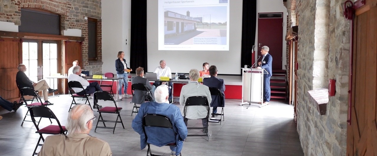 In einem Saal wird ein Vortrag mit einer Päsentation auf einer Leinwand gehalten. Der Redner steht rechts vorne an einem Rednerpult.
