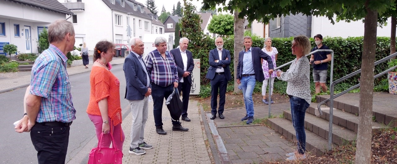 Staatssekretärin Milz steht im Kreis mit anderen Personen, während eine Dame etwas vorträgt. Sie befinden sich draußen auf einem Bürgersteig vor einem Treppenaufgang, beidseitig mit Geländern.