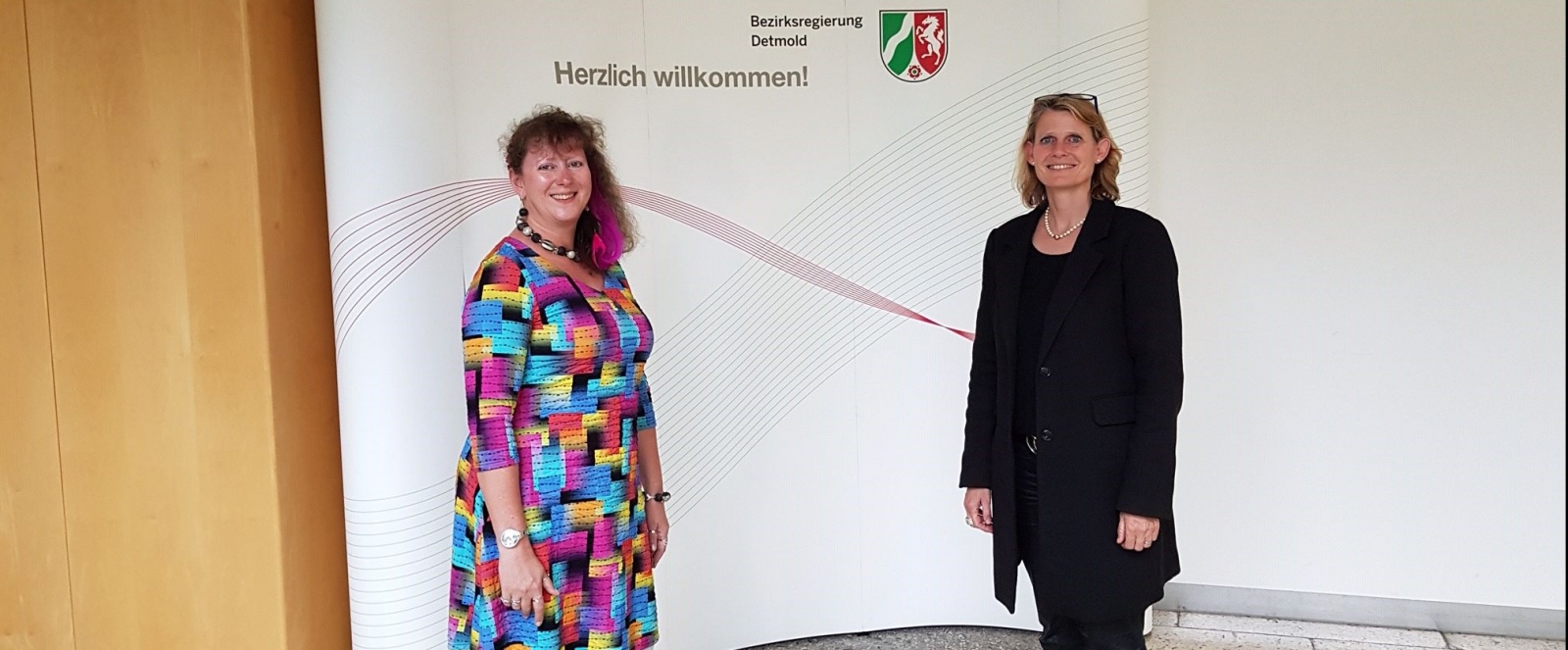 Staatssekretärin Andrea Milz im bunten Kleid steht mit der Bezirksregierungspräsidentin Judith Pirscher vor einer weißen Plakatwand mit der Aufschrift "Herzlich Willkommen!" und darüber in kleiner Schrift "Bezirksregierung Betmold" mit dem NRW-Wappen.