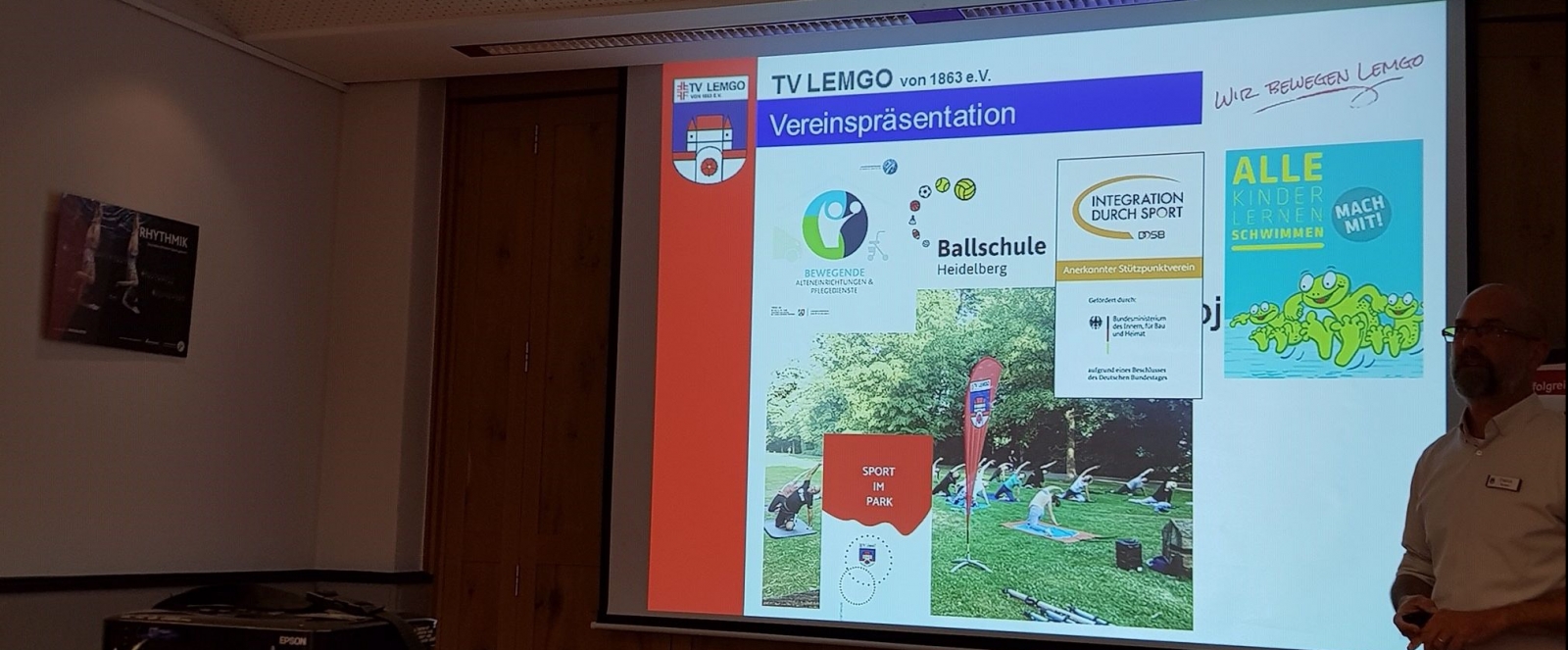 Das Foto zeigt einen Raum mit einer sehr großen Bildschirmpräsentation von TV Lemgo e.V. an einer Wand. Rechts davon steht ein Mann mit Brille und weißem Hemd im Halbschatten.