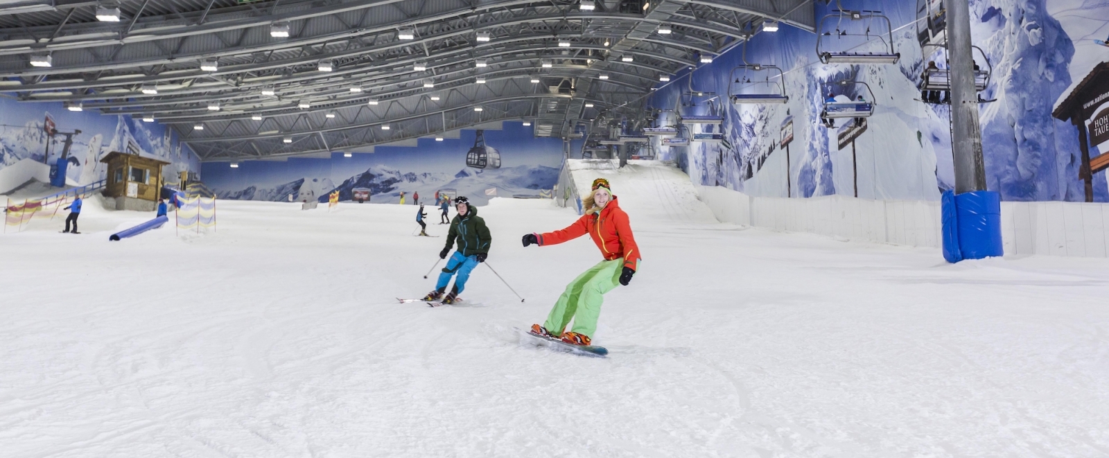 Eine Snowboardfahrerin und ein Skifahrer fahren in einer Skihalle eine Piste hinunter. Rechtsseitig bringt ein Sessellift die Skifahrer nach oben. 