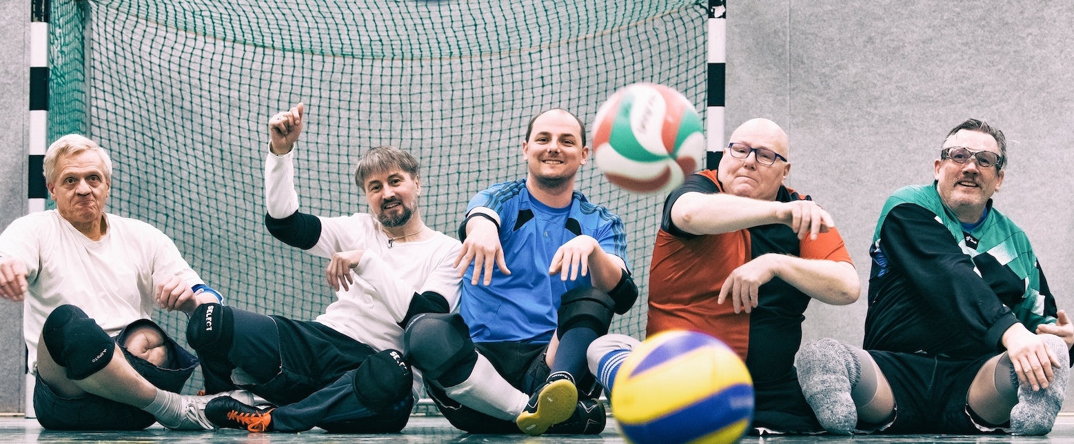 5 Herren spielen Sitzball. Hinter ihnen ein Tor, vor ihnen 4 bunte Bälle.