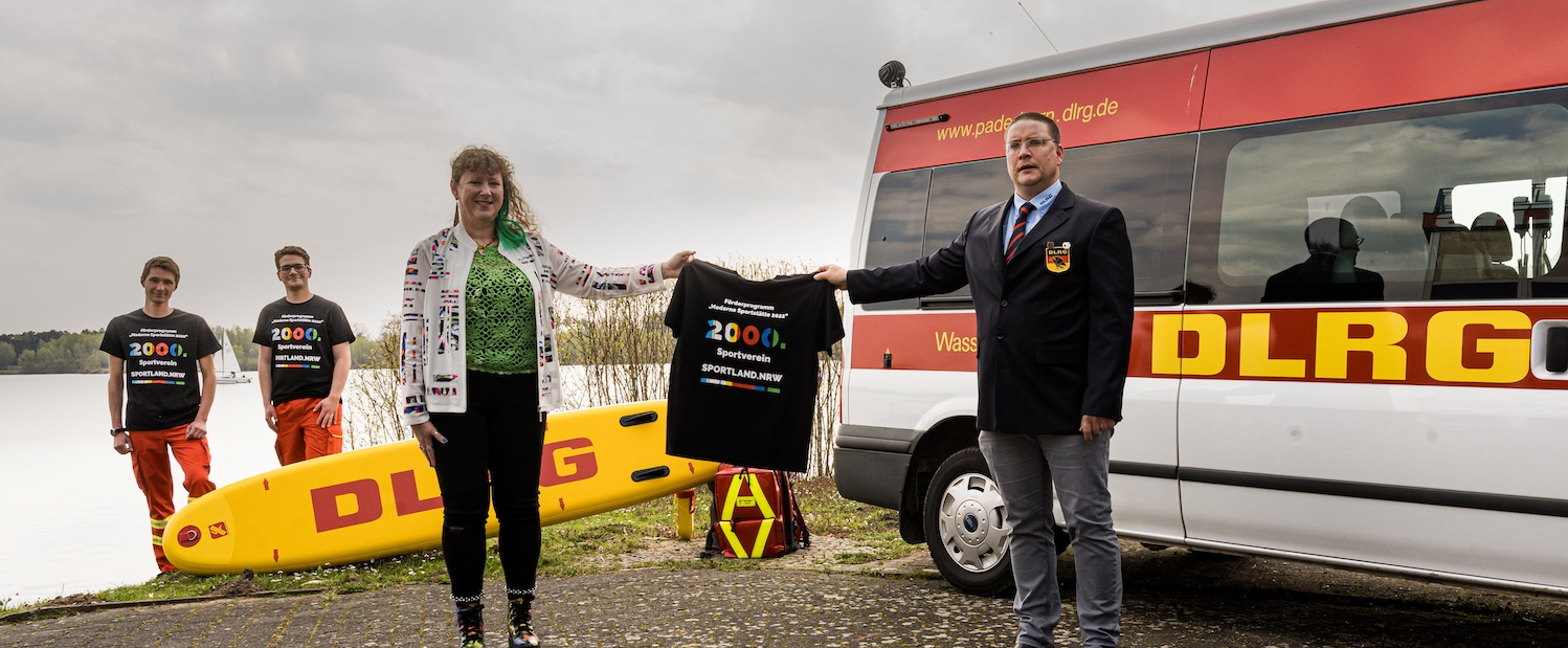 Ein Mann und eine Frau halten ein Tshirt hoch, im Hintergrund stehen zwei weitere Männer neben einem Rettungswagen.