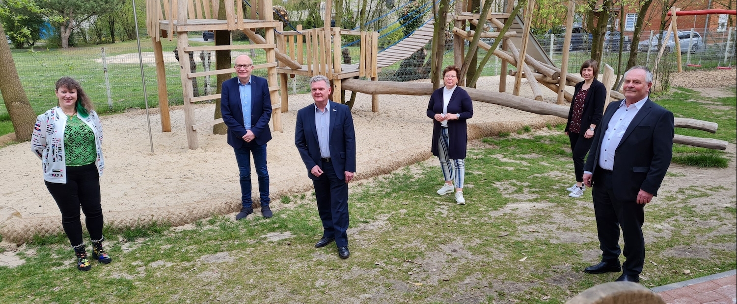 Staatssekretärin Andrea Milz steht mit 5 weiteren Menschen auf einer Wiese vor einem Sandspielplatz mit vielen Holzgerüsten.