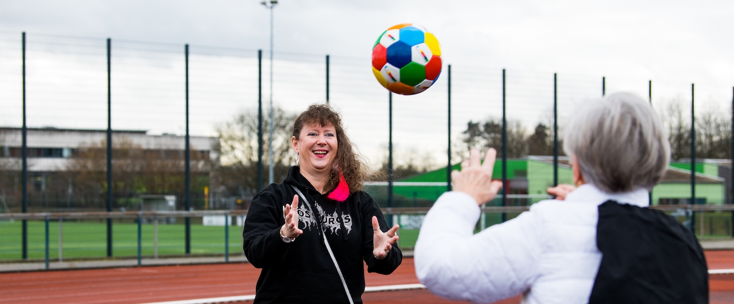 Frau Erika Rischko in weißer Daunenjacke und schwarzer Hose wirft der Staatsekretärin Andrea Milz in schwarzen Sportbekleidung einen bunten Sportland.NRW-Fußball zu. Die sechseckigen Felder des Fußballs haben die Sportland.NRW-Farben Weiß, Rot, Gelb, Grün, Blau und Dunkelblau. Auf dem Rücken tragt Frau Rischko einen schwarzen Sportland.NRW-Rucksack mit Sportland.NRW-Logo unten auf der Frontseite.