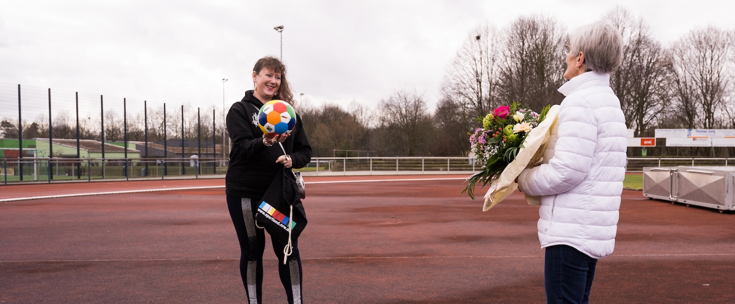 Frau Erika Rischko in weißer Daunenjacke und schwarzer Hose und Schuhen hält einen Blumenstrauß in ihren Händen. Ihr gegenüber stehend hält Staatsekretärin Andrea Milz in schwarzer Sportland-NRW-Sportbekleidung einen bunten Sportland.NRW-Fußball und einen Sportland.NRW-Rucksack in ihren Händen. Die sechseckigen Felder des Fußballs haben die Sportland.NRW-Farben Weiß, Rot, Gelb, Grün, Blau und Dunkelblau. Sie stehen auf einem Sportplatz.