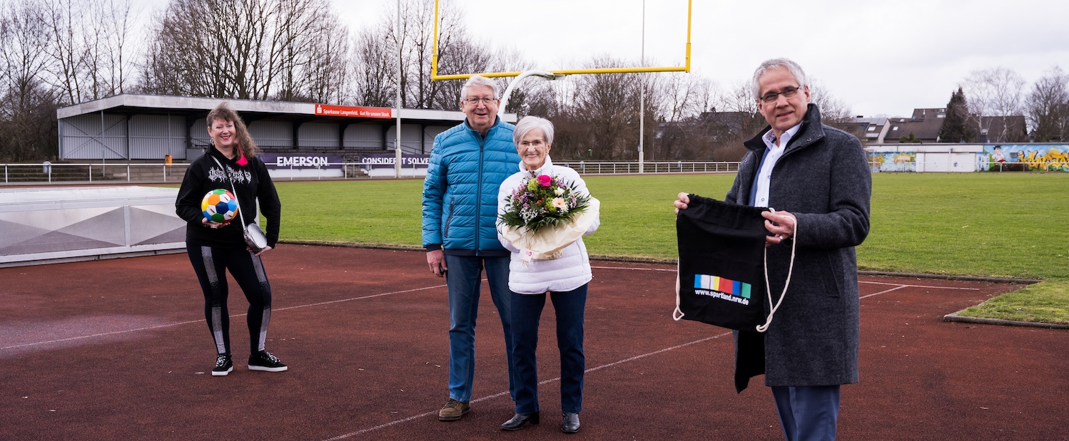 Frau Erika Rischko in weißer Daunenjacke und schwarzer Hose und Schuhen hält einen Blumenstrauß in ihren Händen. Neben ihr steht ein Herr in blauer Jacke. Links steh die Staatsekretärin Andrea Milz und ganz rechts der Bürgermeister von Langenfeld.