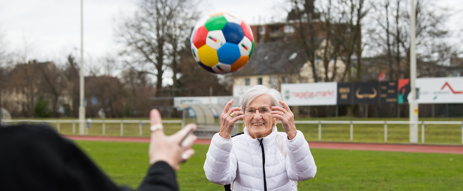 Frau Erika Rischko in weißer Daunenjacke und schwarzer Hose wirft der Staatsekretärin Andrea Milz in schwarzen Sportbekleidung einen bunten Sportland.NRW-Fußball zu. Die sechseckigen Felder des Fußballs haben die Sportland.NRW-Farben Weiß, Rot, Gelb, Grün, Blau und Dunkelblau.