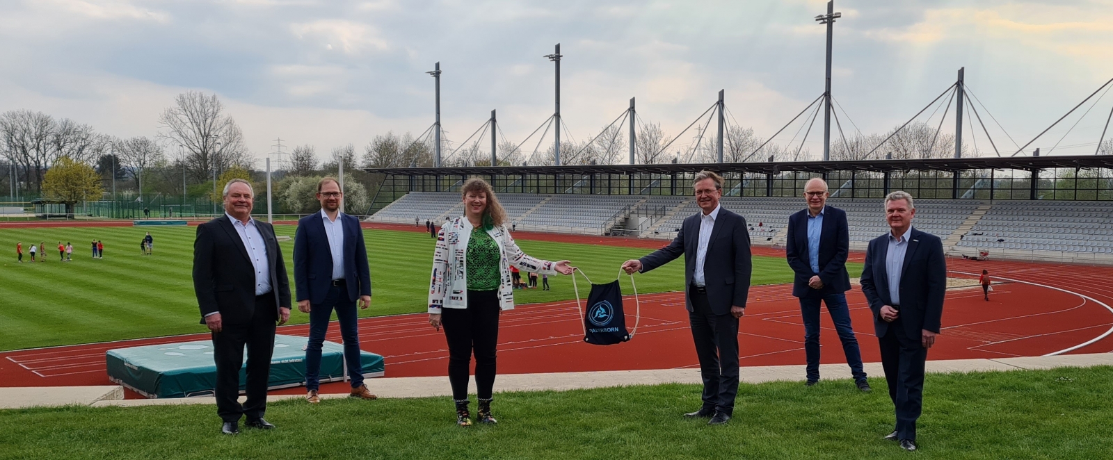 Staatssekretärin Andrea Milz steht mit 5 weiteren Menschen vor einer Sportstätte mit Tartanbahnen und einem Rasenplatz in der Mitte.