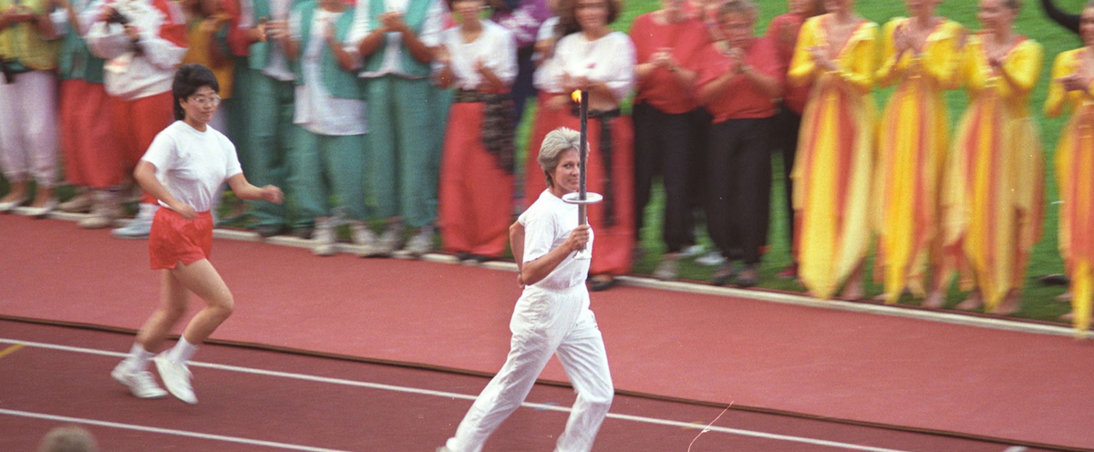 Eine Frau in weißer Kleidung läuft mit einer brennenden Fackel in der Hand über eine rote Laufbahn