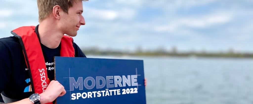 Ein junger Mann sitzt auf einem Schlauchboot und hält ein blaues Schild 