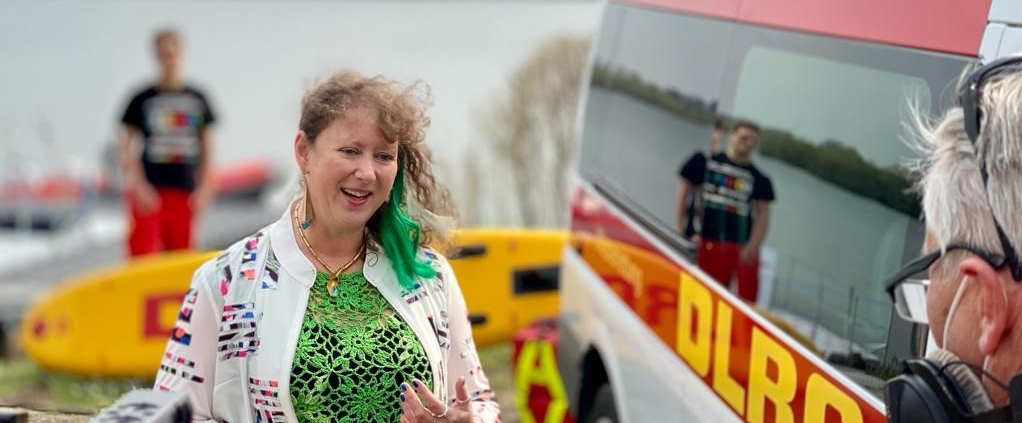 Eine Frau spricht vor einer Kamera, im Hintergrund ist ein Rettungswagen des DLRG