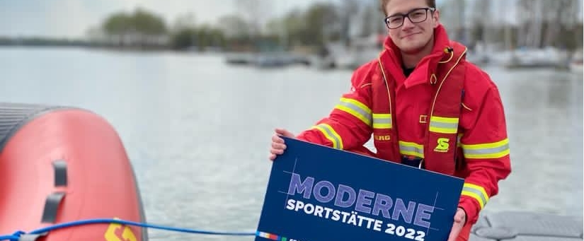 Ein Mann kniet auf einem Steg neben einem Schlauchboot und hält ein blaues Schild 