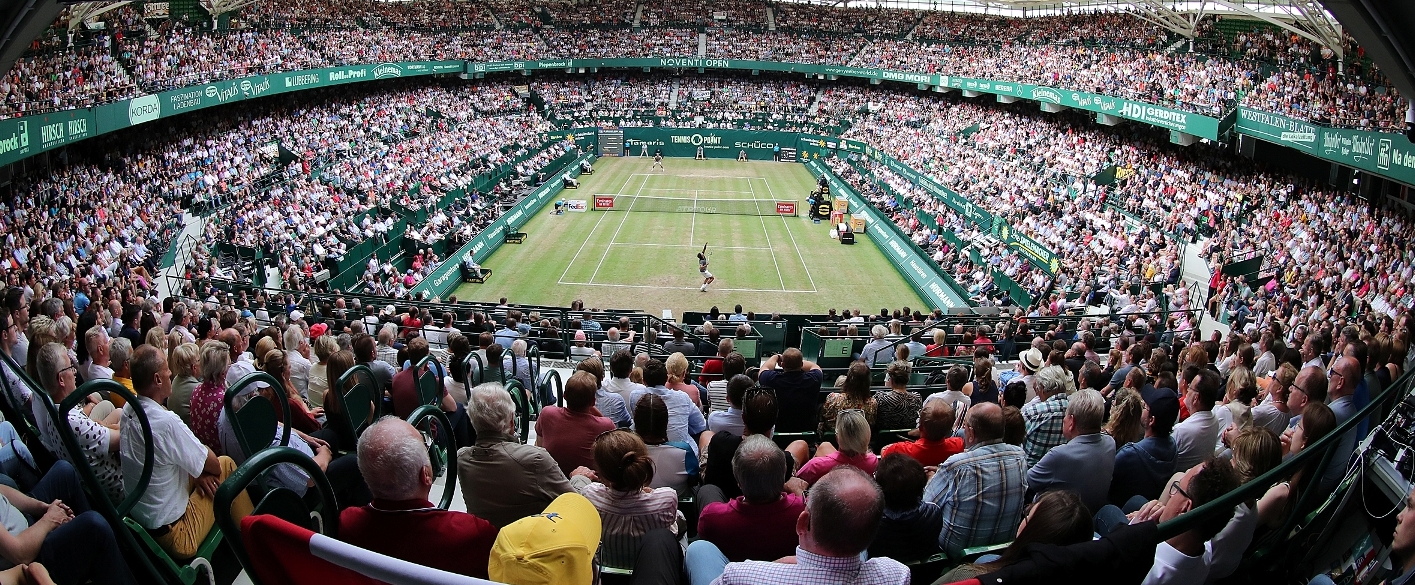 Tennis Arena in Fischaugenperspektive. Auf dem Feld spielen zwei Tennisspieler, während das ausverkaufte Stadion gespannt zuschaut.