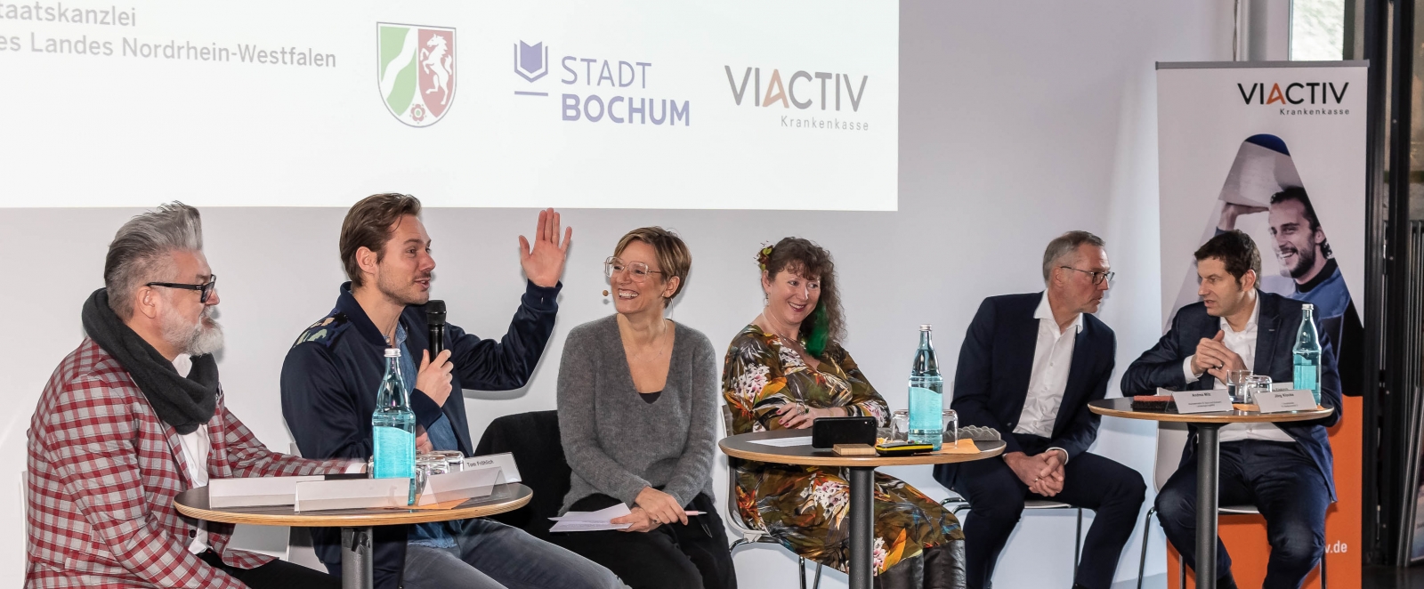 Staatssekretärin Milz sitzt mit mehreren Personen auf Barhockern mit Rückenlehne, vor sich befindne sich runde Stehtische. Ein Herr spricht gerade in ein Mikrofon.