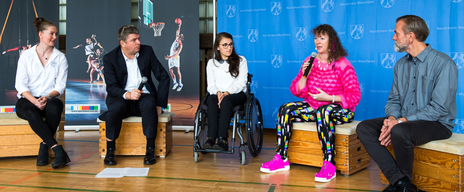 Staatssekretärin Milz sitzt mit vier Personen in einer Sporthalle, hinter ihnen mehree Plakatwände mit NRW-Logos.