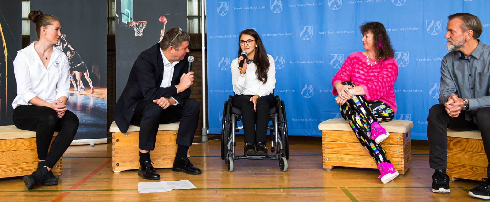 Staatssekretärin Milz sitzt mit vier Personen in eienr Sporthalle. Ein Herr spricht gerade mit einer Sportlerin im Rollstuhl, beide halten ein Mikrofon in einer Hand.