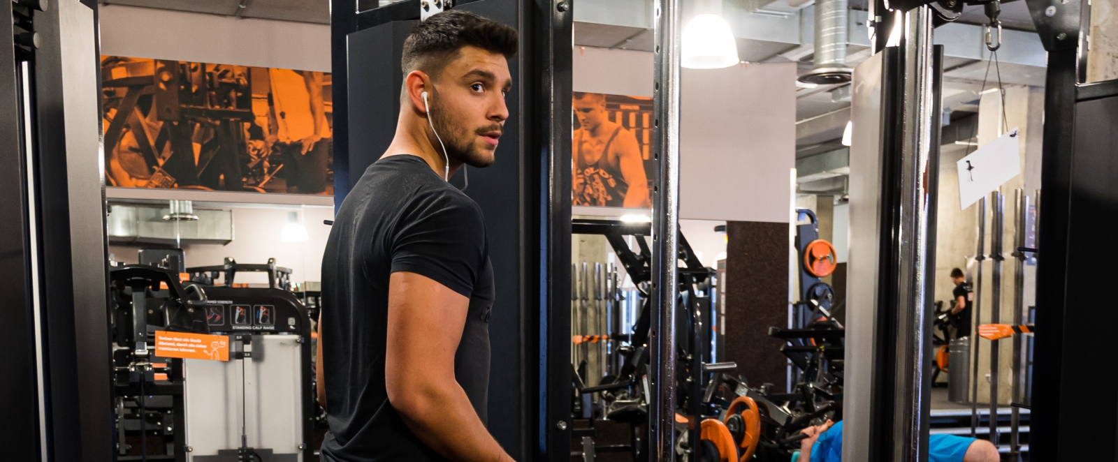 Ein junger Mann in schwarzen T-Shirt und Bermudahose mit weißen Socken und Sportschuhen geht durch einen Fitnessraum.