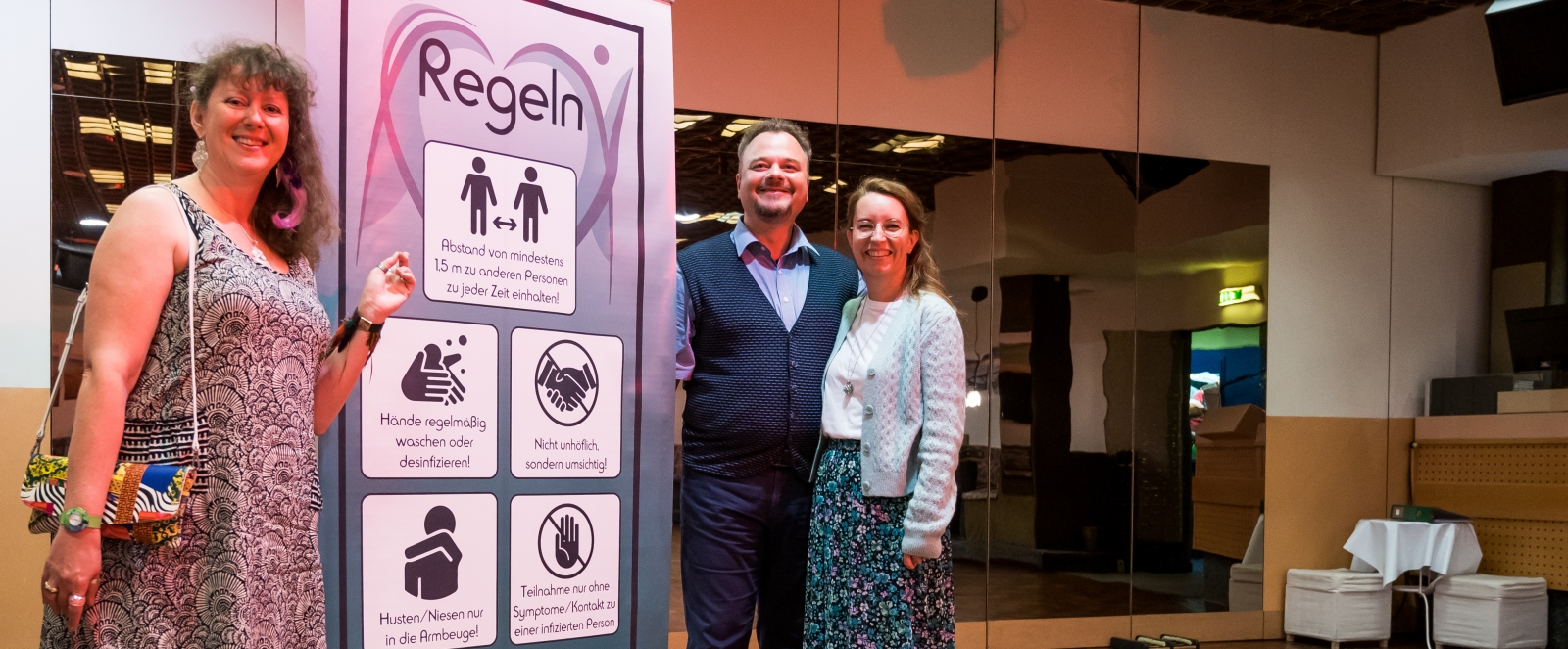 Staatssekretärin Milz steht mit einem Paar an der Hygiene-Plakatwand des Tanzhauses.