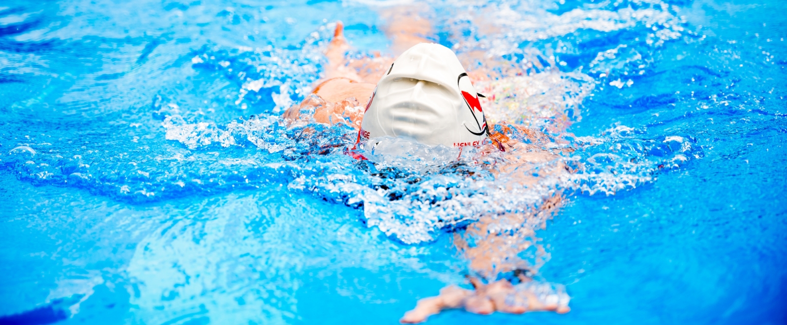 In einem Schwimmbekcken schwimmt ein Schwimmer mit weißer Badekappe im Kraulstil auf den Betrachter zu, sein Gesicht ist unter Wasser, sein linker Arm  ist nach vorne gestreckt unter Wasser.