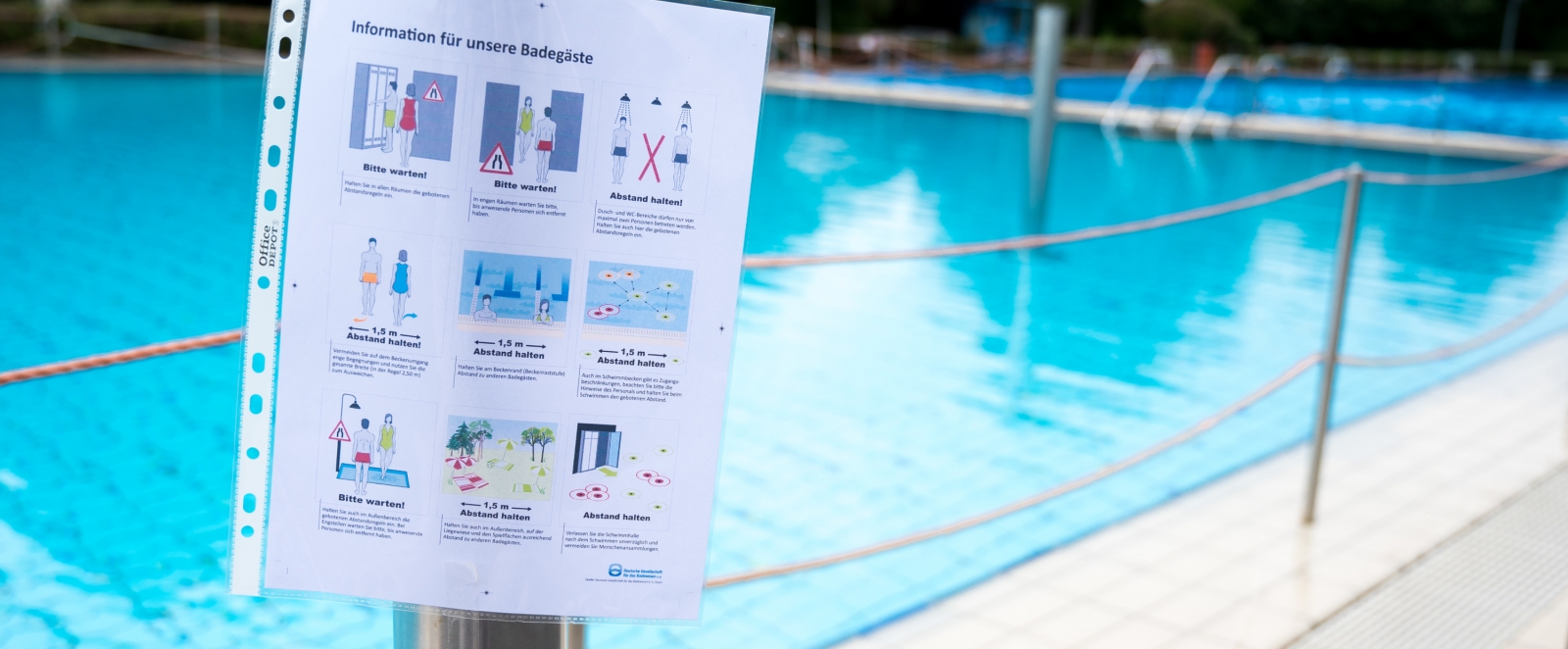 Hygiene-Hinweise in einer Prospekthülle an einem silbernen Mast neben einem offenen Schwimmbecken.