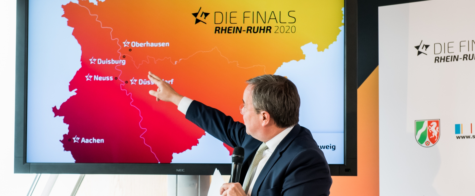 MInisterpräsident Laschet steht vor einer Bildschirmpräsentation und zeigt dabei auf einer NRW-Karte in Richtung Ruhrgebiet.