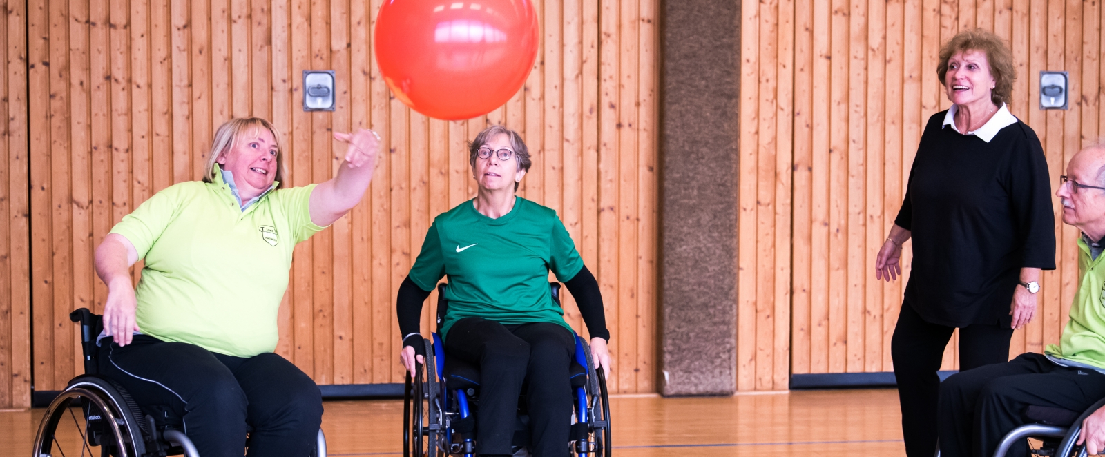 Zwei Sportlerinnen und ein Sportler jeweils in Rollstühlen sitzend spielen mit einem großen Ball. Eine schwarz bekleidete Dame steht lächelnd dabei und sieht zu.