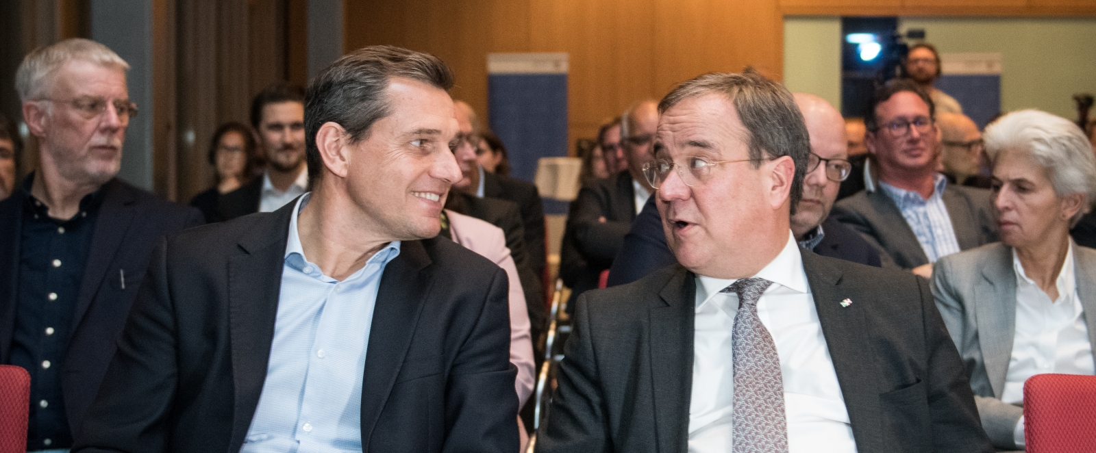 MInisterpräsident Laschet sitz im Publikum und spricht mit einem Herren.