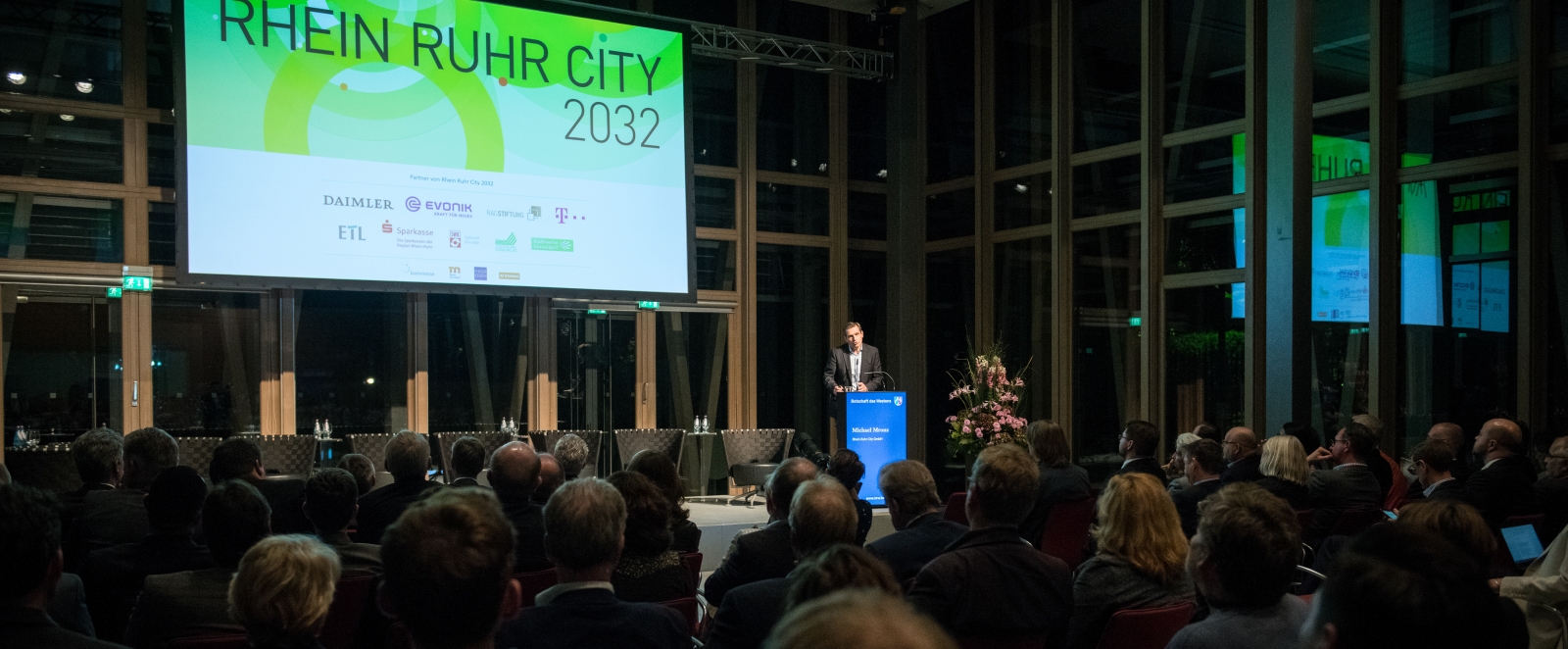 MP Laschet hält eine Rede vor einem großen Publikum in einem Saal. Vorne oben hängt eine Leinwand mit einer Präsentation auf der steht auf grünem Grund: Rhein Ruhr City 2032.