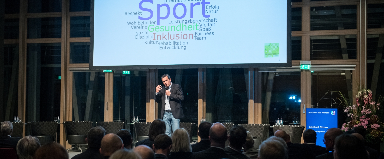 Ein Mann hält eine Rede vor einem großen Publikum in einem Saal. Vorne oben hängt eine Leinwand mit einer Präsentation auf der steen auf blauen Grund einzelne Wörter wie: Sport, Bildung, Integration, Inklusion, Gesundheit, etc.