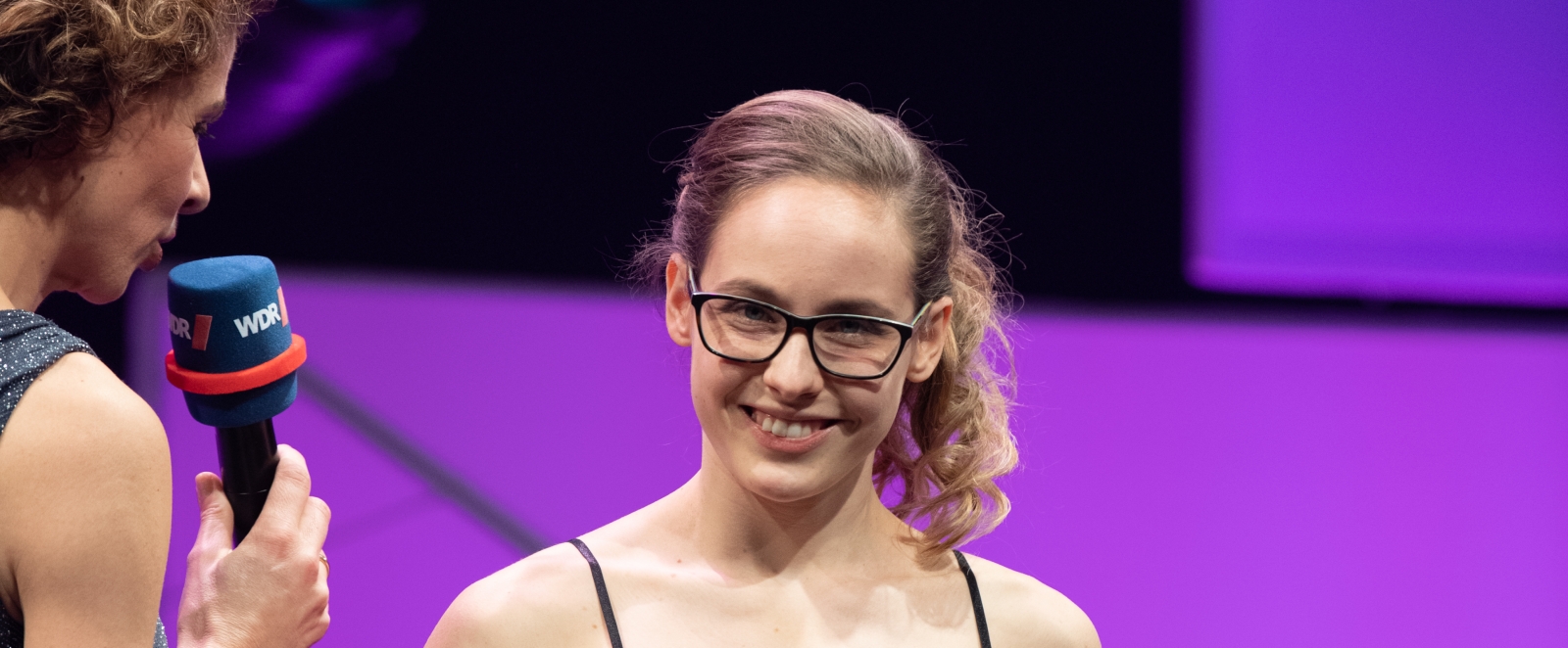 Eine Sportlerin steht im schwarzen ärmellosen Abendkleid und hält den Felix-Award in ihren Händen. Links neben ihr steht die Moderatorin mit einem Mikrofon in der Hand.
