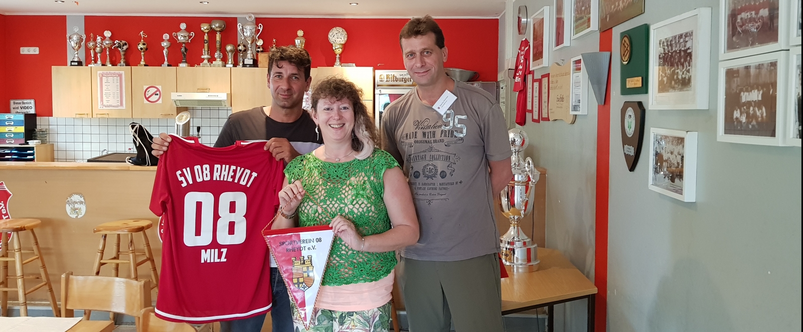 Staatssekretärin Andrea Milz mit zwei Vereinsmitgliedern im Clubraum, neben sich ein roten Vereinstrikot. Hinten an der Rückwand stehen viele Pokale und rechts an der Wand mehrere eingerahmte Fotos.