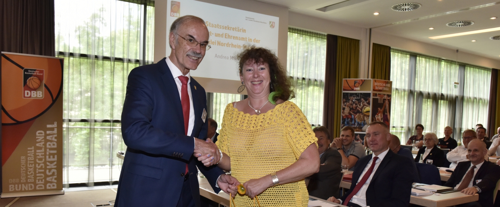 Staatssekretärin Andrea Milz im gelben Oberteil steht mit einem Herrnin einem Saal.  Im HIntergrund sitzen viele Gäste in mehreren Reihen .