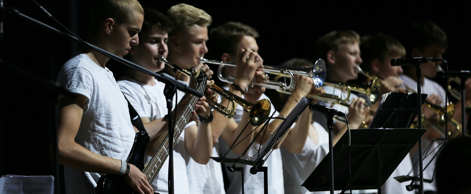 Musiker spielen stehend auf Gitarren und Blasinstrumenten. Sie sind in weißen T-Shirts und in blauen oder schwarzen Jeanshosen gekleidet. Vor sich stehen Standmikrofone.