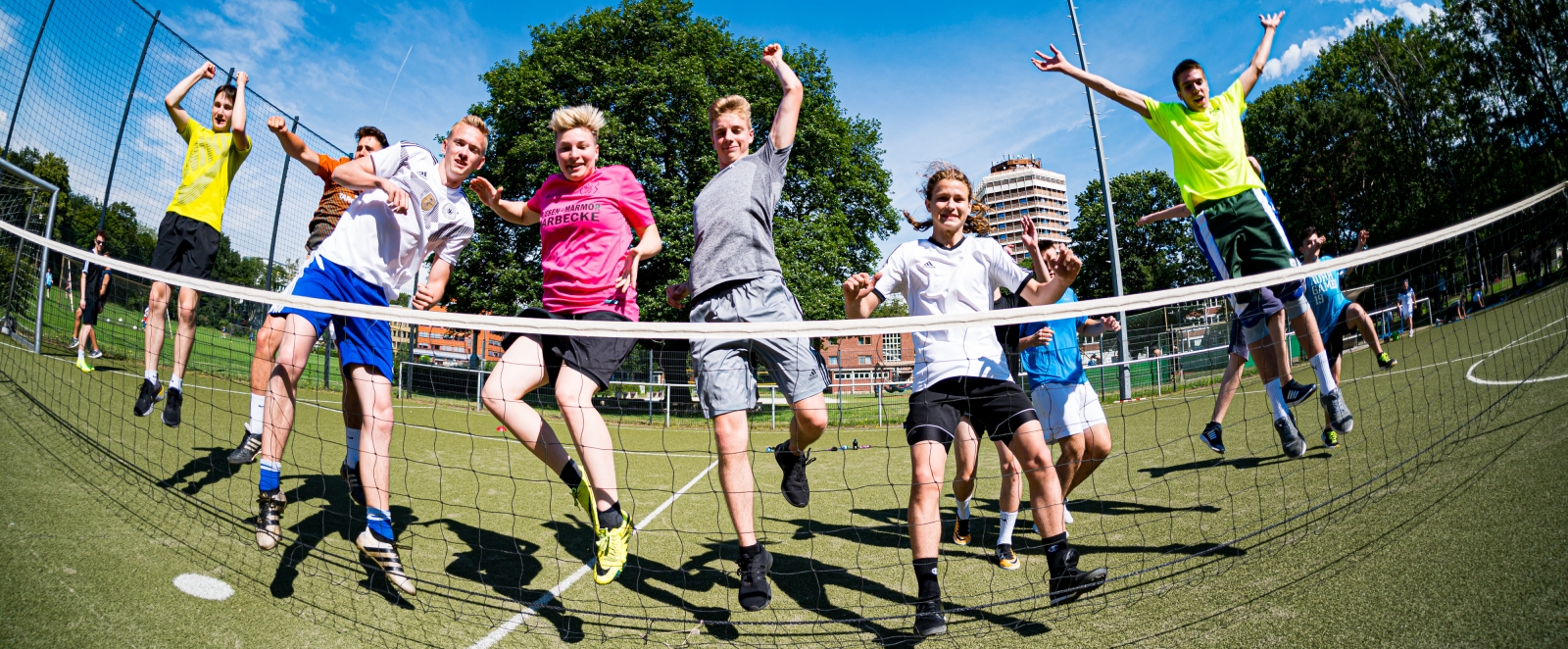 Mehrere Jugendliche in Sportkleidung springen vor Fruede mit hochgerissenen Armen vor einem Badminton-Netz in die Höhe.