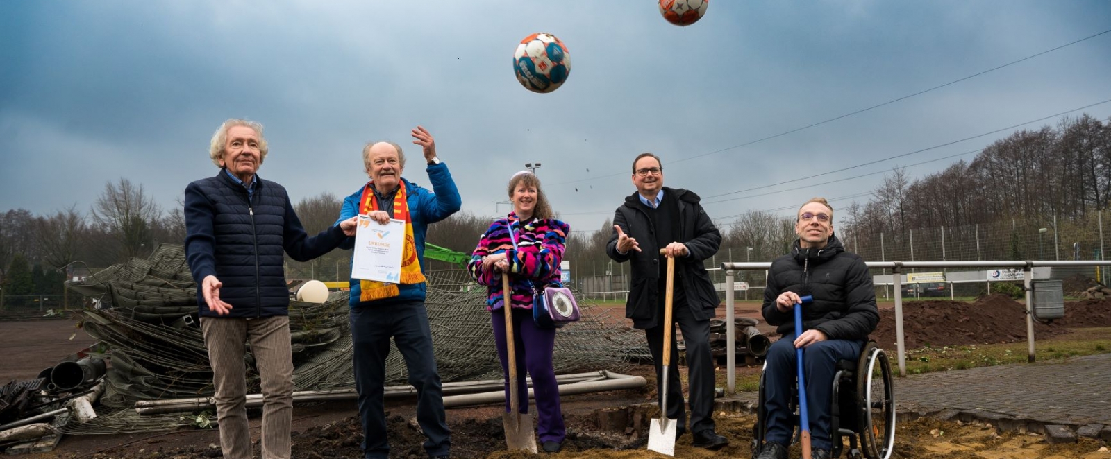 Staatssekretärin Milz steht mit vier Männern auf einem Bauplatz für den ersten Spatenstich, ein Mann sitzt im Rollstuhl. Alle haben einen Spaten in der Hand. Zwei Sportland.nrw-Fußbälle sind hoch in die Luft geworfen worden.