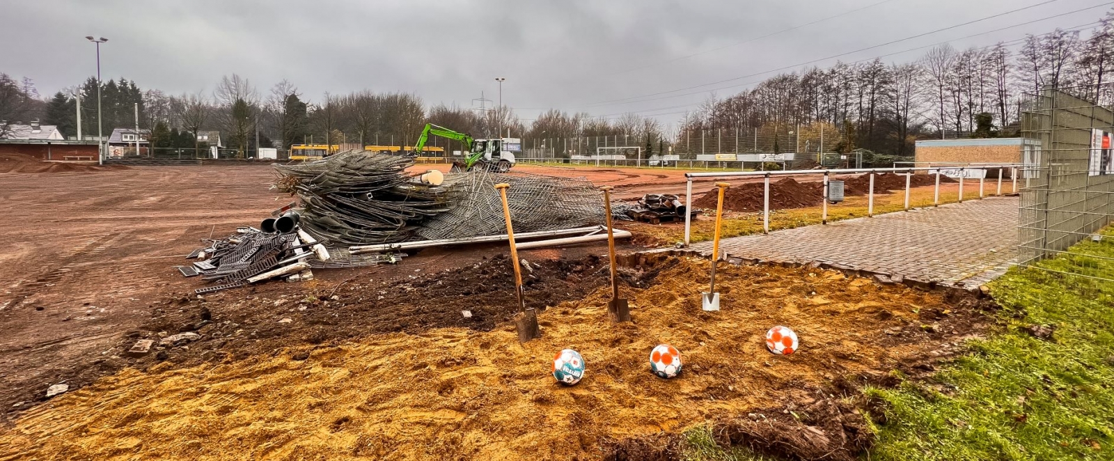 Man blickt über eiinen abgerissenen Sportplatz, der Rasen ist entfernt. Vorne rechts im Bild sieht man drei Spaten, die aufrecht in den Sand gesteckt wurden, vor jedem Spaten liegt ein Sportland.nrw-Fußball.