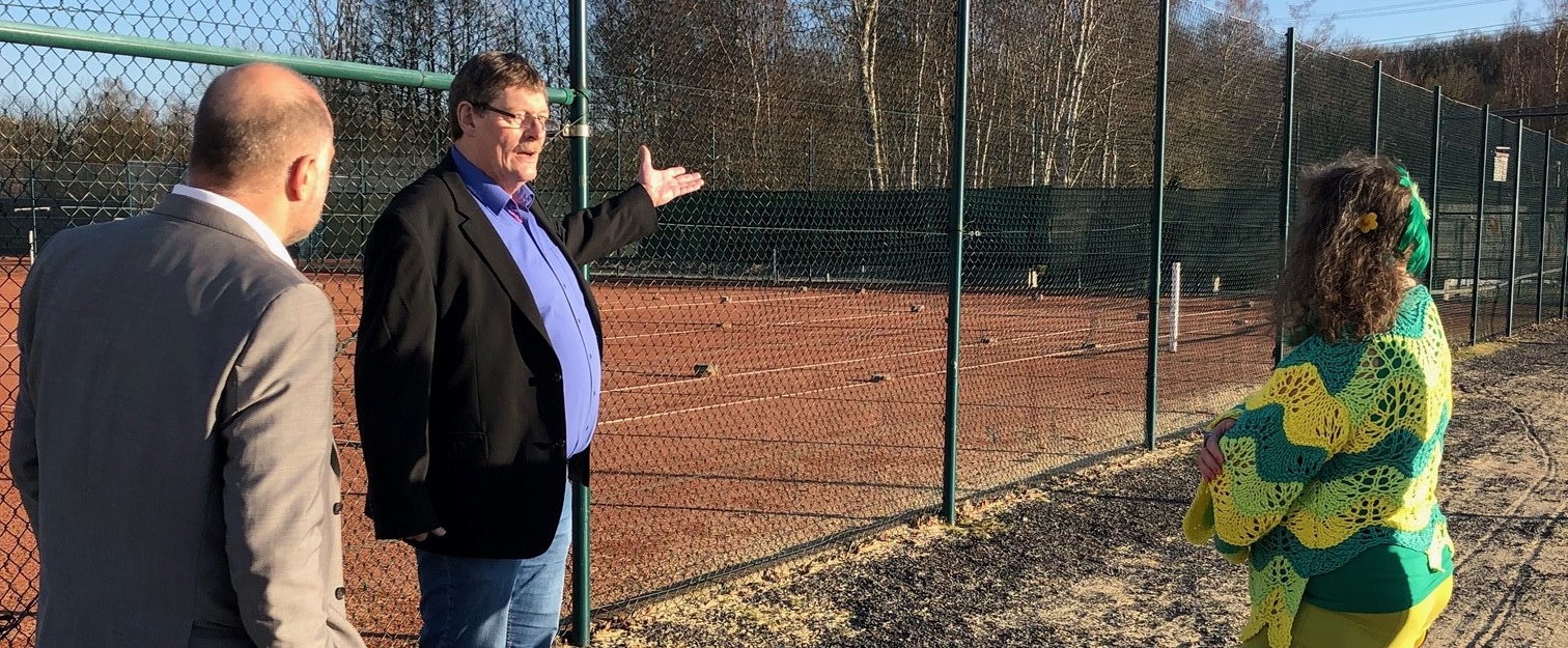 Staatssekretärin Milz steht mit zwei Herren vor einem eingezäunten Tennisplatz.