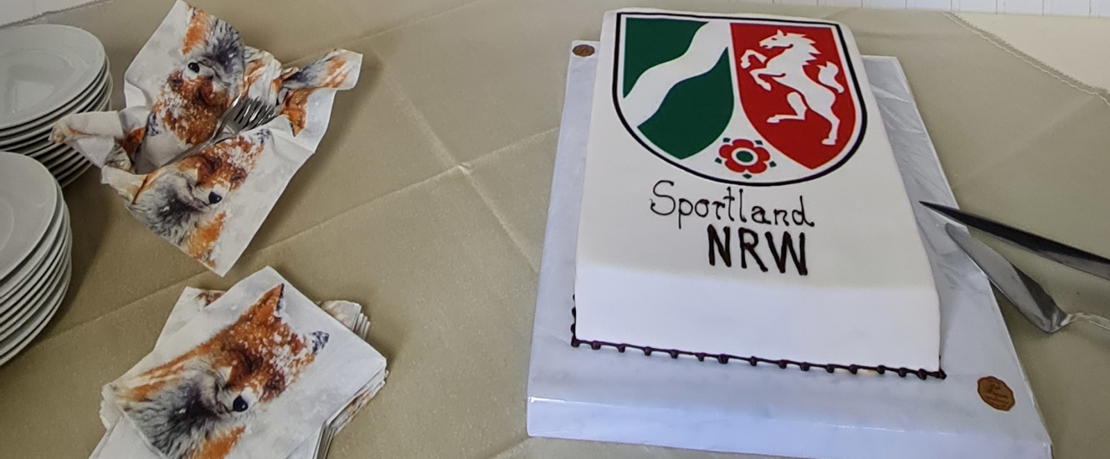 Ein flacher, quaderförmiger Kuchen mit weißer Glasur und einem großen NRW-Wappen (rot, grün und weiß) darauf. Unter dem Wappen steht noch "Sportland NRW". Rechts sieht man einige Teller und bunte Servietten. An der Wand dahinter hängen mehrere, kleine, gezeichnete Hundebilder in schwarzen Rahmen.