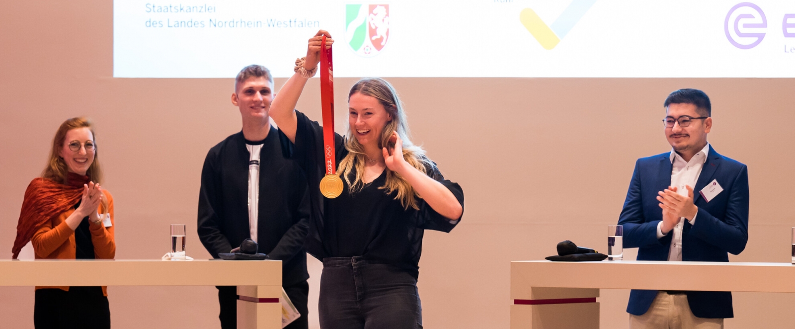 Eine Frau mit blond-braunen Haaren mit schwarzen Top und schwarzer Hose steht auf der Bühne und hält lächelnd eine goldfarbene Medaille an einem roten Band hoch.
