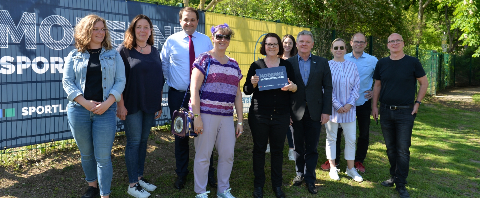 Gruppenbild mit Staatssekretärin Milz beim Turn-Verein-Ehrenfeld mit dem Schild "Moderne Sportstätte 2022".