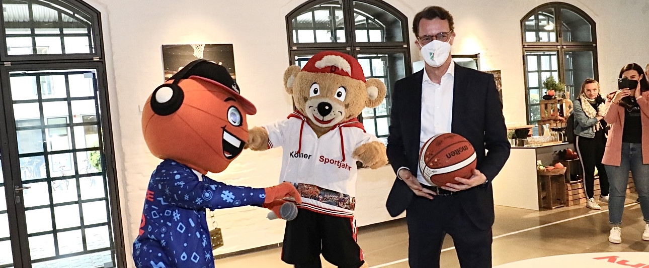 Hendrik Wüst und Maskottchen in Sporthalle mit Basketball