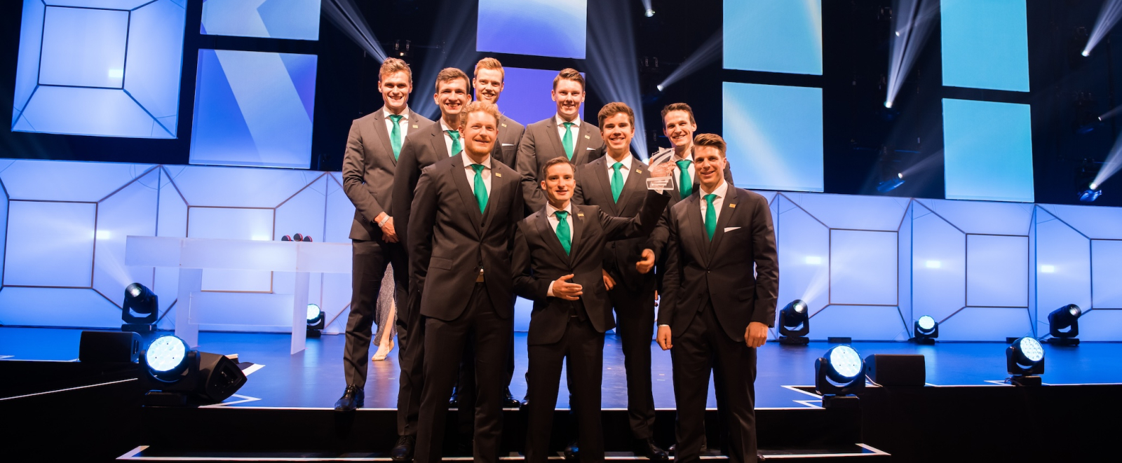 Neun Männer in dunkelgrauen Anzügen und grünen Krawatten stehen auf einer Treppe, die zur Bühne hinaufführt.