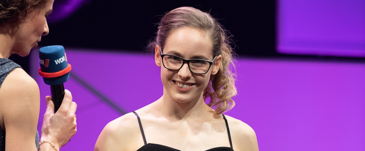 Eine Sportlerin steht im schwarzen ärmellosen Abendkleid und hält den Felix-Award in ihren Händen. Links neben ihr steht die Moderatorin mit einem Mikrofon in der Hand.