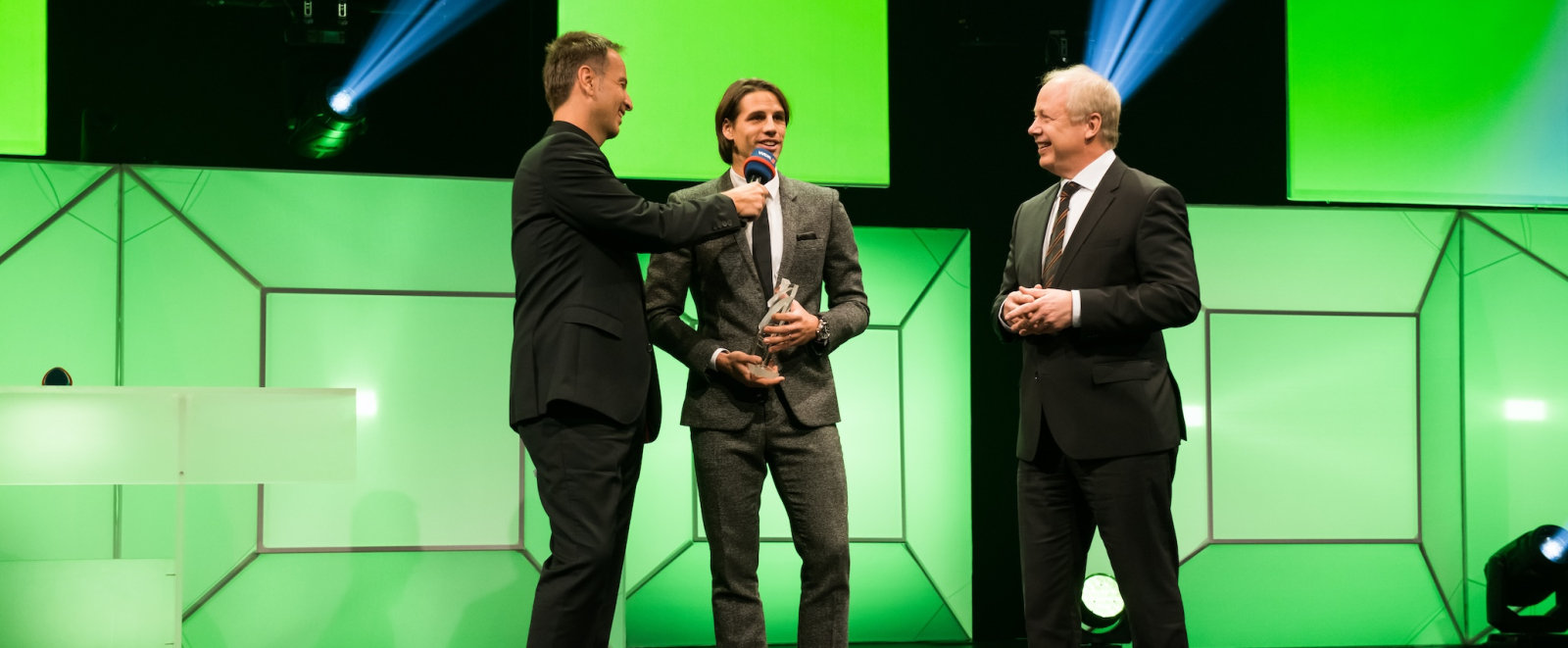 Drei Männer in Anzügen stehen auf einer Bühne, der Hintergrund ist grünlich