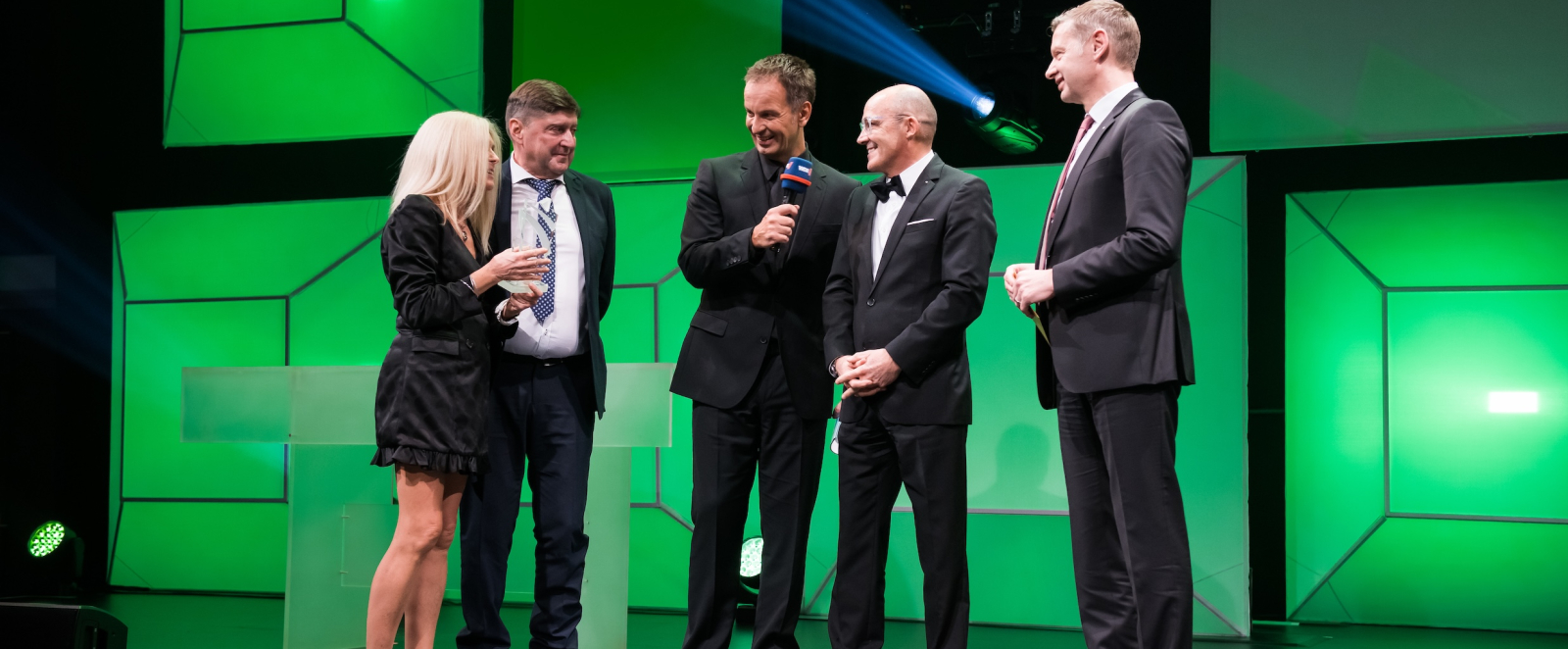 Eine Dame und vier Herren stehen auf einer Bühne, der Hintergrund ist grünlich 