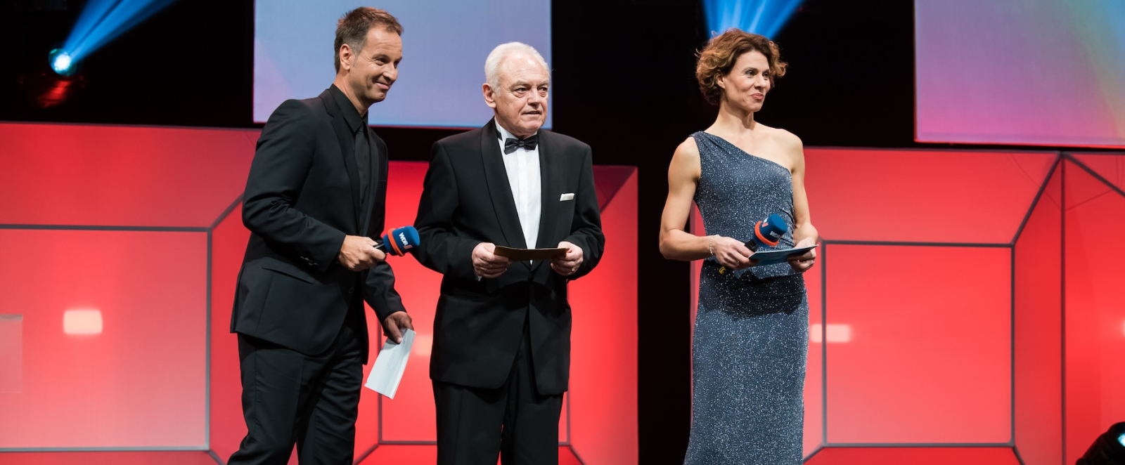 Zwei Herren und die Moderatorin stehen auf der Bühne, der Hintergrund ist rötlich beleuchtet.